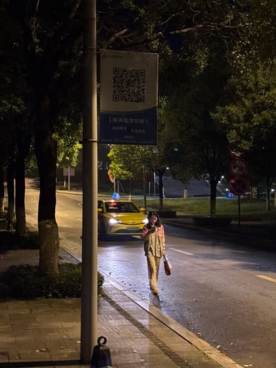 The image shows an urban scene at night in the Nan'an District of China. A person, wearing a plaid shirt and tan pants, lies prone on the wet pavement near a curb. A yellow vehicle with headlights illuminated is adjacent to the person on the road. A sign with a QR code is attached to a utility pole along the sidewalk. Trees are visible in the background. In the distance, traffic signs are present. The environment appears to be wet, possibly after rain. The exact actions or interactions are not clear from the image alone.
