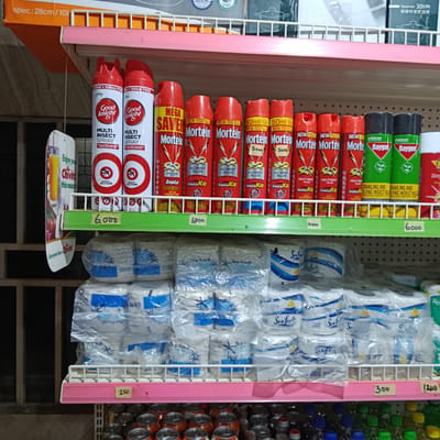 The image displays a well-stocked retail shelf inside a store located in Gwarinpa, Nigeria. The scene is devoid of people.
The uppermost shelf in the background, partially visible, contains packaged items, possibly kitchen appliances, indicated by text such as "Steamer 30cm" and "spec: 28cm / 10L."
The primary focus is the middle shelf, which is divided into two layers. The upper layer showcases various aerosol insect sprays. From left to right, there are two red and white "Good Knight Multi Insect Spray" cans, labeled as "600ml + 100ml FREE" and featuring an "Instant Action" mosquito logo. Adjacent to these are multiple red "Mortein" cans, with various labels including "Mega Saver," "New Improved Formulation," "All Insect Killer," and "InstaKill." Some Mortein cans have handwritten price tags of "5000." Further to the right are two green and black "Baygon Crawling and Flying Insect Killer" cans by SC Johnson. Price tags placed directly below these sprays read "6,000," "6,500," "4,000," and "6,000."
The lower layer of the middle shelf contains numerous individually plastic-wrapped packages of "Sea Fresh" brand paper products, likely toilet paper or paper towels, distinguished by white packaging and blue decorative elements. Below this section, price tags of "250," "300," and "1,200" are visible.
The bottom shelf, partially visible at the very bottom of the frame, holds rows of aluminum beverage cans, predominantly with orange/red tops, and an assortment of plastic bottles with liquids, capped in various colors like green, blue, yellow, and red.
A promotional cardboard hanger for "Chivita" is suspended on the left, displaying the phrases "Enjoy your Chivita this season" and "Everyone has a Chivita," along with an image of a drink in a glass. The background on the far left reveals a dark, gridded structure, possibly a window with security bars. The shelving units are constructed of white wire with pink shelf dividers.