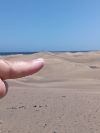 The image depicts a desert landscape with sand dunes under a clear blue sky and a visible body of water in the distance. A human finger is in the foreground, pointing towards the distant dunes. Two small figures stand on top of the farthest dune visible. The ground in the foreground is sandy with small dark objects. The location is likely the Maspalomas Dunes in San Bartolomé de Tirajana, Spain, given the characteristic desert landscape next to the sea.