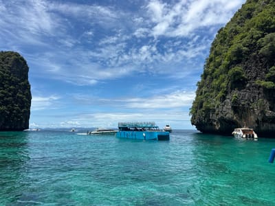 The image captures a scenic view of the turquoise waters off the coast of Ao Nang, Thailand, a popular tourist destination. In the foreground, the crystal-clear water exhibits a vibrant greenish-blue hue, with gentle ripples indicating calm sea conditions. Several boats are visible in the scene. A prominent blue boat with a multi-level structure and a canopy is positioned in the mid-ground, appearing to be a passenger ferry or tour boat. To its left, a white speedboat with the name "NATTHICHA" is partially visible. Further in the distance, other boats, including speedboats and potentially a larger vessel, are dotted across the horizon. The background is dominated by towering limestone cliffs, characteristic of the Phi Phi Islands, which are often visited from Ao Nang. These cliffs are covered in lush green vegetation, adding to the tropical ambiance. The sky above is a bright blue, interspersed with scattered white clouds, suggesting fair weather. The overall scene portrays a serene and picturesque maritime landscape, typical of the islands accessible from Ao Nang.
