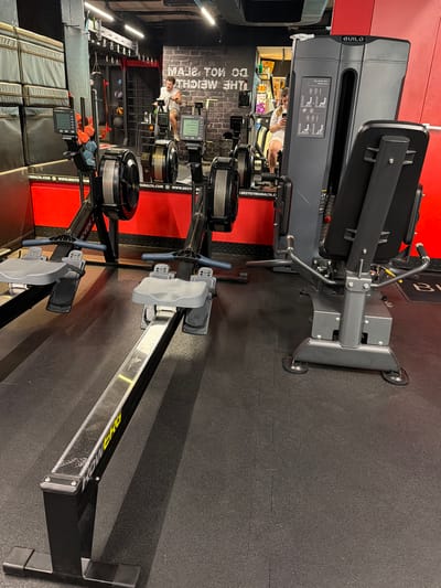 This image captures an indoor gym environment in Saint Paul's Bay, Malta, illuminated by bright overhead lights. The floor is covered with black rubber mats, and the walls feature sections of red and brick-patterned finishes, along with reflective surfaces in the background.
The foreground and midground are dominated by exercise equipment. Multiple Concept2 Rowerg rowing machines are visible, with the closest one clearly showing its "ROWERG" branding in yellow on the rail, along with its seat, footrests, and display screen which reads "Concept2".
Further back, a grey "BUILD" brand hip abduction and adduction machine is prominent. It features an instructional sticker with diagrams and the text "Hip Abduction Adduction", demonstrating its proper use. In the reflection of a large mirror or reflective wall behind this machine, a man wearing a light-colored t-shirt is seen interacting with gym equipment, and another person is partially visible. Additional gym apparatus, including weight racks and possibly a punching bag, are discernible in the deeper background reflections. Snacks and water bottles are also visible on a counter.
Text visible on the walls, often reversed due to reflections, includes "MAJE TOI", "THE MEICH", and web addresses such as "SMALTA.COM", "WWW.BESTGYMSMALTA.COM", and "I.BESTGYMSMALTA.C", confirming the gym's location in Malta. The scene depicts a functional fitness space, likely active during the day.