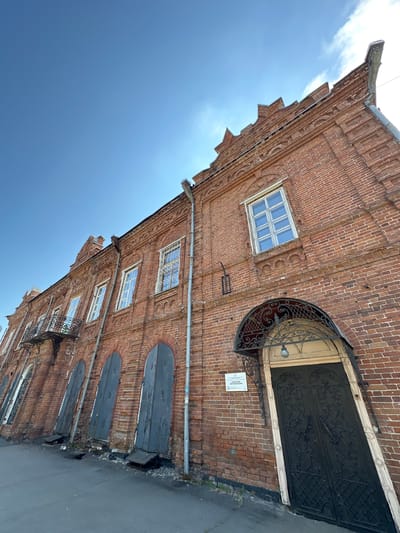 This is a low-angle, exterior, wide shot of a red brick building on a bright, clear day in Biysk, Russia. The building features decorative brickwork along its facade, a prominent cornice with stepped gables, and several arched windows and doorways. On the left side, there are three large, grey, weathered wooden doors with small openings at their bases. To the right of these, a black metal double door is set within an arched entrance, topped by an ornate black metal awning. A sign reading "СИБИРСКИЙ ТОРГОВЫЙ БАНК" (Siberian Commercial Bank) is visible to the left of the entrance. A balcony with intricate black metal railings juts out from the second story on the left side of the building. Drainage pipes run vertically down the brickwork. The ground in front of the building is paved with asphalt. The sky is a clear, vibrant blue with a few wisps of clouds.