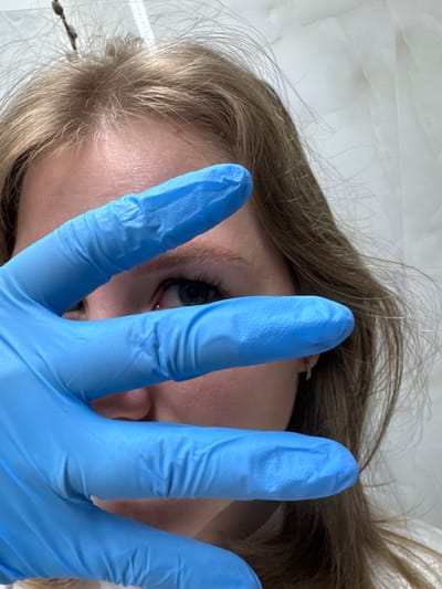 A medium, low-angle shot depicts a person with blond hair, fair skin, and blue eyes partially obscured by a hand wearing a blue latex glove. The glove's fingers are spread across the person's face, obscuring most of their features. The background appears to be a white or light-colored surface, possibly a wall or ceiling with a subtle texture. The location is not discernible.