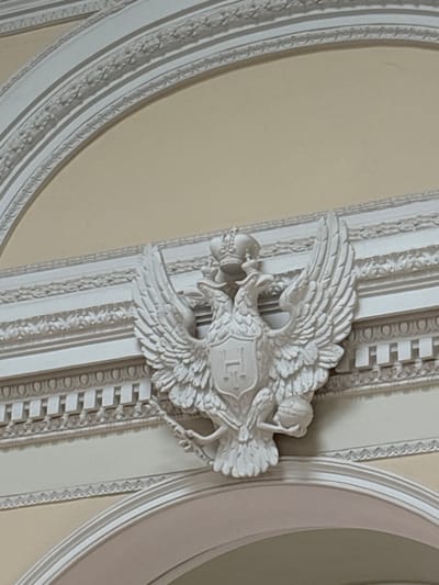 The image shows a white stucco bas-relief of a double-headed eagle, a symbol of the Russian Empire, adorning an interior wall. The eagle is depicted with its wings spread, wearing a crown, and clutching a scepter and an orb in its talons. A shield bearing the letter "H" is centered on its chest. The eagle is positioned above a decorative frieze with a pattern of circles and rectangular motifs. The background wall is painted a pale yellow. The bas-relief is part of the architectural ornamentation of the Grand Palace in Gatchina, Russia, specifically located within the Pavlovsk Hall or a similar grand reception room. The overall scene suggests a historical and opulent setting.