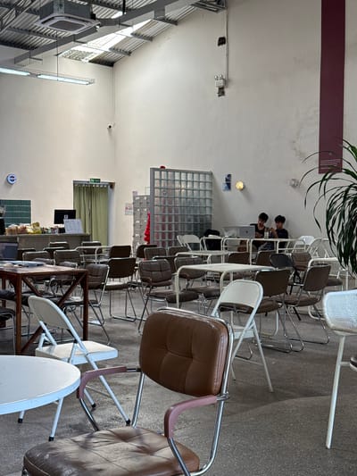 This is a photograph of the interior of a cafe with an industrial aesthetic, located in Tam Kỳ, Vietnam. The cafe features a high, corrugated metal ceiling with exposed beams and ductwork. Natural light filters in through skylights. The walls are painted a light, neutral color and show some signs of wear.
In the foreground, a brown leather armchair with chrome legs and pink armrests is prominent. To its left is a round, white table. Further into the cafe, rows of tables and chairs are arranged. Many of the chairs have brown leather seats and backs with chrome frames. There are also white folding chairs visible.
In the middle ground, a counter area is visible with a laptop, what appear to be baked goods, and other cafe items. A "TOILET" sign hangs above a doorway draped with a green curtain. Behind the counter, there are lockers and a partition made of glass blocks.
In the background, two young males are seated at a table with a laptop, suggesting they are working or studying. The overall ambiance is casual and utilitarian. The specific location within Tam Kỳ, Vietnam, is not discernible from the image itself.