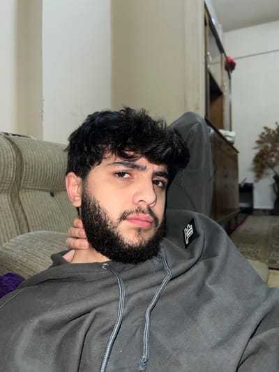 A person with dark, curly hair and a full beard is centered in the image, looking at the camera. They are wearing a gray hoodie. The background is blurred, showing a light-colored wall and furniture. The image is rotated, with the person's left ear near the bottom left corner. Their right hand is visible, touching their neck.