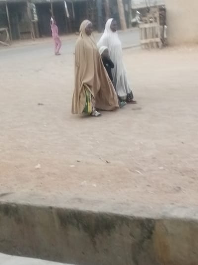 Two women in traditional clothing walk down a dusty street. One woman wears a light brown hijab and robe, and the other wears white. A small child is walking between the women. In the background, a person in pink clothing walks across the street. Buildings are visible in the background on either side of the road.