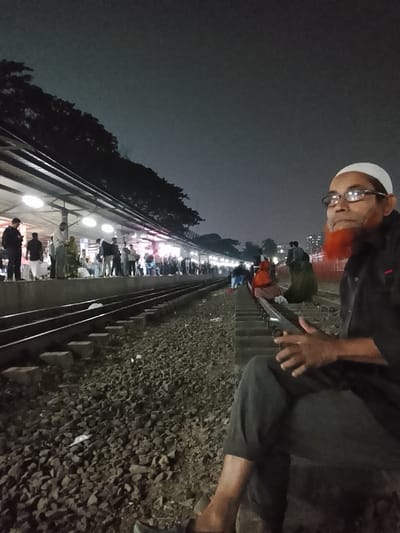 The image depicts a bustling railway station at night, likely in Shibpur, Bangladesh. In the foreground on the right, a man with a white cap (kufi), glasses, and a distinct reddish-orange beard is seated directly on the ground beside the railway tracks. He is wearing dark attire and holds a smartphone with both hands, looking towards the platform with a contemplative expression.
Two parallel railway tracks traverse the midground, with sleepers and gravel ballast clearly visible. Further along the tracks to the right, several other individuals are also seated on the ground, seemingly waiting.
On the left side of the frame, a well-lit platform stretches into the distance, bustling with a crowd of people. They stand under a canopy illuminated by numerous overhead lights. The platform appears busy, suggesting people are waiting for a train. Above the platform and trees are silhouetted against a dark, cloudy night sky. Scattered debris and litter are visible on the railway ballast. The scene captures a typical late-night waiting environment at a South Asian train station. No text is visible in the image.