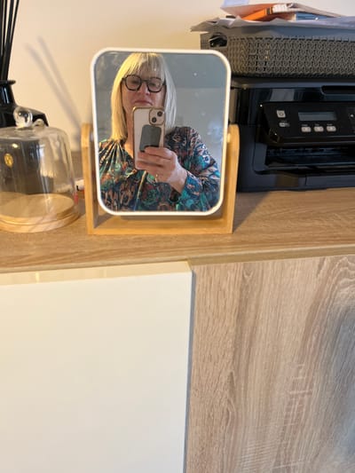 The image depicts an indoor scene in Chambœuf, France. A blonde woman, wearing dark-rimmed glasses and a multicolored patterned blouse, is reflected in a rectangular mirror with rounded corners, framed in light wood. She holds a light-colored smartphone with a dark rectangular attachment on its back in her right hand, positioned as if taking a photograph of her reflection. A ring is visible on her left ring finger.
The mirror rests on a light wood-grain surface. To the left of the mirror, a tall, slender black diffuser bottle with black reeds and a clear glass cloche with a wooden base are present. To the right of the mirror, a black HP multifunction printer is visible, topped by a dark grey woven plastic basket filled with various papers and an orange folder. Below the light wood-grain surface, a white cabinet door is visible on the left, while the wood-grain pattern continues on the right side of the lower portion of the image. The background wall is a plain, light cream color.