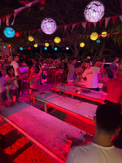 The image depicts an outdoor evening gathering, likely a party or event, illuminated by colorful spherical lanterns and string lights, creating a vibrant and festive atmosphere. The scene is bathed in red and pink neon lighting. In the foreground, several people are visible, some engaged in what appears to be a drinking game involving cups placed on long tables. A woman in the middle ground is actively participating, holding a cup and looking towards others. In the background, a bar area is set up, with a bartender serving drinks. The setting seems to be a beach or a casual outdoor venue with trees overhead. The overall impression is one of lively social interaction and celebration. The location is identified as Unknown, Indonesia.