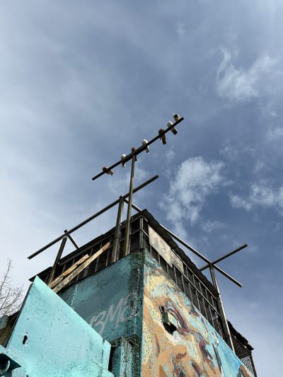 The image shows a view looking up at a dilapidated structure, likely an abandoned building or observation tower, against a cloudy blue sky. Several pigeons are perched on a horizontal metal bar above the structure. The building itself is a multi-level concrete structure with peeling paint in blue and yellow, adorned with graffiti. Metal railings and fencing are visible, as well as what appear to be wooden planks. The scene suggests an urban environment, specifically in Lyubertsy, Russia.