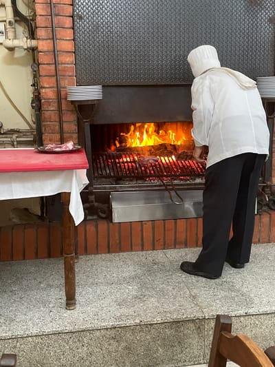 In Mogadouro, Portugal, an indoor scene depicts a person, viewed from behind, in a white chef's jacket, hat, and dark trousers, actively grilling meat. The individual stands before a large, built-in brick and metal grill containing vigorous orange flames under a metal grate where multiple pieces of meat are cooking. The person uses a pair of long, silver metal tongs to handle the meat. To the left of the grill, a small wooden table draped with a white tablecloth features a red top surface, upon which a dark plate holds two raw, thick-cut steaks with bone. Above the grill, a section of textured dark metal cladding is visible, with a small stack of white plates resting on a narrow shelf attached to it. The floor consists of light grey speckled tiles, with a raised step in the foreground. Exposed utility pipes are visible along the brick wall in the upper left background.