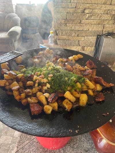 The image depicts an outdoor or semi-enclosed cooking scene in Petrivka, Ukraine. In the foreground, a large, black, circular metal cooking pan (likely a *saj*) rests on a red metal stand. The pan is filled with actively cooking food, emitting steam. The primary food items are diced, golden-brown fried potatoes and pieces of fried meat, some with crispy, darkened surfaces. Centrally placed on top of the cooked food is a mound of fresh, chopped green herbs, including dill and segmented spring onions (scallions) with light pinkish-white bases. Visible oil or fat sizzles around the food. A dark wooden spatula is positioned on the left side of the pan, partially resting on the food.
In the background to the left, a light-colored, upholstered armchair is partially visible, behind which a blurred human figure in dark clothing is discernable amidst rising steam or haze. To the right background, a wall constructed from horizontal courses of light brown, rough-hewn stone blocks forms a textured backdrop. A clear plastic water bottle with a yellow cap sits on a surface near this wall. Adjacent to the bottle and the wall, a silver-colored metallic appliance with black controls and vents is partially visible. The cooking setup rests on a speckled granite-like surface, transitioning to a tiled floor in the lower left. A dark brown ceramic pot is partially visible beneath the cooking pan on the right side.
