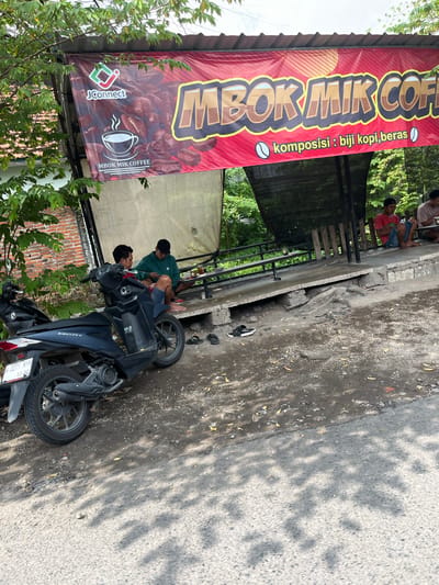 In the city of Jati Alun Alun, East Java, a banner with the text "JConnect" and "MBOK MIK COFFEE" is displayed above a coffee stall. The banner features a stylized coffee cup logo and mentions "komposisi: biji kopi, beras". Two young men are seated on a bench at the stall, one wearing a green hoodie and the other a red shirt. A black motorcycle is parked in front of the stall. The ground is a mix of dirt and pavement, with some fallen leaves scattered around. The area is partially shaded by trees and overhead cover. In the background, there is a brick wall and dense vegetation. A second motorcycle is visible behind the first one. Several pairs of sandals are on the ground near the bench.