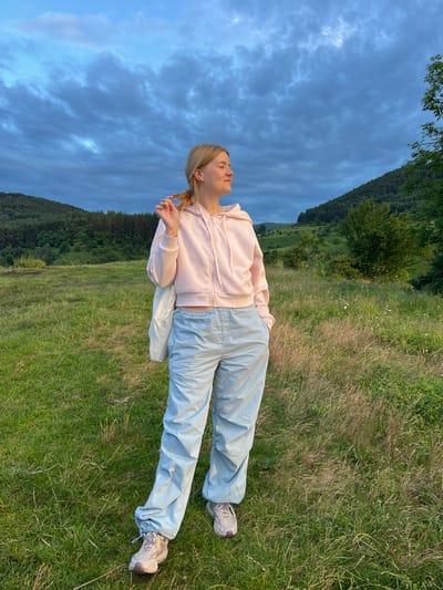 A young Caucasian woman with blonde hair pulled into a braid stands in a grassy, hilly landscape under a cloudy blue sky. She is wearing a light pink hooded sweatshirt and light blue baggy pants. She holds a strand of her hair with her right hand and has her left hand in her pocket. A white bag is slung over her shoulder. The background consists of rolling green hills covered in trees and vegetation. The scene is located in Kislovodsk, Russia.