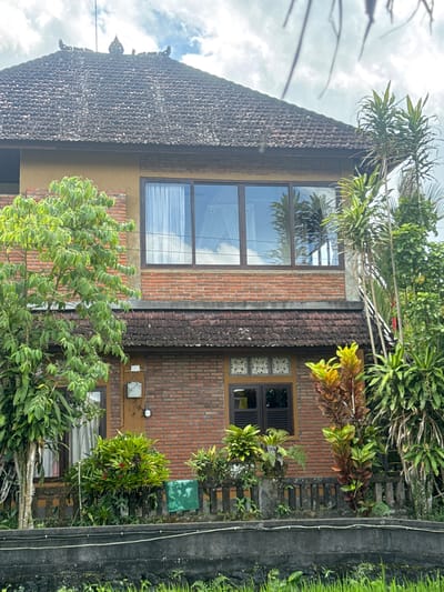 The image depicts a two-story building constructed with red brick and yellow stucco, situated in Tegallalang, Indonesia. The building features a tiled, sloping roof with decorative elements at its apex. Large windows are present on both floors, with the upper floor having a row of three expansive windows that reflect the sky and surrounding greenery. The lower floor has several smaller windows, some with wooden shutters, and an exterior electrical box. The building is surrounded by lush vegetation, including trees and various plants, some of which are growing near a decorative stone fence. The foreground shows a rough concrete barrier and dense green grass, suggesting proximity to a natural landscape. The overall impression is of a tropical dwelling integrated with its environment.