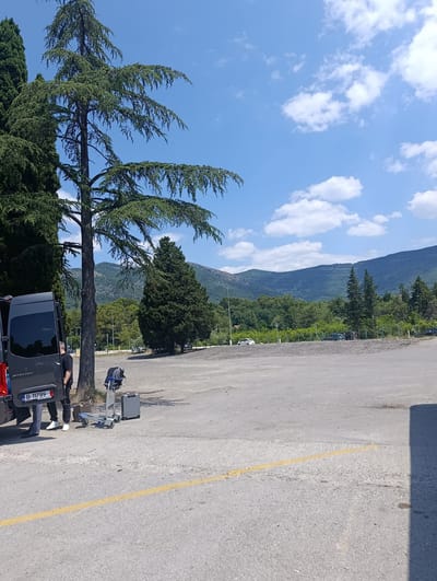 An outdoor daytime scene in Mrčevac, Montenegro, shows a parking lot with several vehicles and a group of people near a dark gray van. The van's rear doors are open, and a man wearing dark clothing and white sneakers stands near it. Beside the van, a luggage cart is placed near two suitcases, one light gray and another brown. Further back, other vehicles are parked, and a line of trees separates the parking lot from a densely forested hillside, topped by a mountain range under a blue sky with scattered white clouds. A yellow line is painted on the asphalt in the foreground.