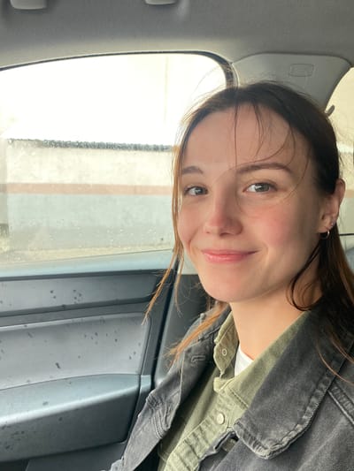 A young woman with brown hair and green eyes smiles at the camera from the passenger seat of a car. She is wearing a green collared shirt under a dark denim jacket. The car's interior is visible, including the door panel and the window. Outside the window, it appears to be raining, with water droplets on the glass and a blurry background suggesting an urban environment. The image was taken in Novokuybyshevsk, Russia.
