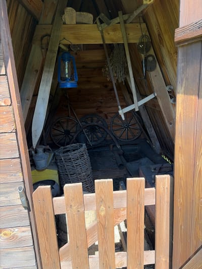 The image depicts the interior of a wooden structure, possibly a shed or storage area, filled with various old tools and equipment. The overall impression is one of disuse and accumulated items.

Prominent objects include:
*   A blue kerosene lantern with a glass bulb.
*   A broom or brush made of natural fibers.
*   Two old wagon wheels, one larger than the other, with spokes.
*   A dark, basket-like woven container.
*   A metal object that resembles an old hand-cranked machine or a piece of agricultural equipment.
*   A wooden plank, possibly part of a tool or a construction element.
*   Two old padlocks, one larger and round, the other smaller and oval.
*   A metal tool with a pointed end, possibly a spear or a pronged implement.
*   A saw blade.

The structure itself is made of weathered wood, with visible beams and planks forming the walls and roof. The lighting suggests it is daytime, with sunlight filtering into the shaded interior, creating dappled light and shadows. Cobwebs are visible in several areas, indicating a lack of recent activity. The location context provided is Kamayevo, Russia. The scene evokes a sense of rural history and the preservation of old tools. There is no visible text in the image.