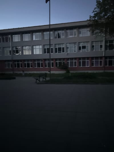 The image captures a modern, multi-story building with numerous windows, possibly a school or office building, in the city of Gorna Malina, Bulgaria. The exterior of the building is predominantly gray and red. A dark flagpole stands tall to the left of the building. In front of the building, a pathway or courtyard is visible, with a dark-colored bench situated on a patch of grass. A person, out of focus and appearing small in the frame, is walking in the distance. The sky is clear, with a gradient from light blue at the top to a darker hue towards the horizon. A portion of a tree is visible on the right side of the frame, its branches casting shadows. The overall scene is dimly lit, suggesting it might be dusk or early evening.