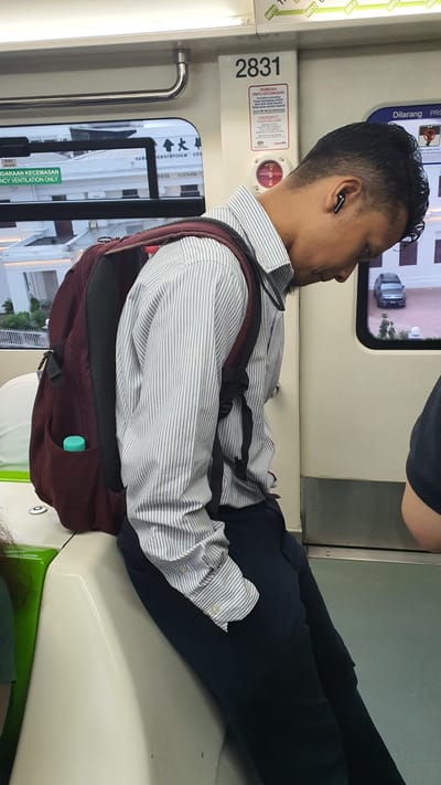 This media file captures a medium shot of a man sitting inside a train, likely in Kuala Lumpur, Malaysia, given the text present. The man is wearing a light blue and white striped dress shirt, dark trousers, and has a maroon backpack slung over his shoulders. He also appears to be wearing wireless earbuds. His legs are crossed, and he is leaning back against the seat. The overall impression is of a commuter traveling.
The setting is clearly a train carriage, indicated by the seats, windows showing passing scenery (including a car outside), and various labels and signage on the train's interior. One prominent sign displays the number "2831" and text in Malay, including "PEMBUKA PINTU KECEMASAN" which translates to "EMERGENCY DOOR RELEASE." Another sign states "DILARANG PRO" (PROHIBITED). Other visible text includes "MR11," "MR10," and "MR9," which could refer to specific train lines or carriage numbers. There are also some Chinese characters visible through the window, suggesting a culturally diverse area.
The lighting suggests it might be daytime, but without further information, the specific time of day and weather conditions cannot be determined. The man's facial expression is neutral, not conveying any strong emotions. The activity is passive; he is simply a passenger traveling. The image is oriented vertically, implying it was taken with the camera held in portrait mode.