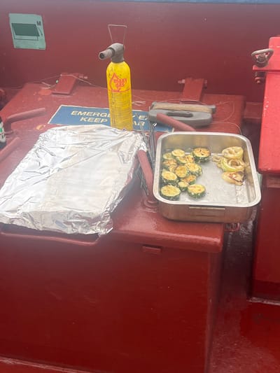 The scene depicts a collection of cooking items and tools on a red-painted metallic surface, likely the deck or superstructure of a vessel, located in Bangor, United Kingdom. In the center, a yellow "maxy gas" blowtorch stands upright. To its left, a rectangular baking tray is fully covered with aluminum foil. To the right of the blowtorch, a silver-colored metal tray contains prepared food, specifically numerous slices of cooked zucchini and several slices of cooked white onion. Behind the foil-covered tray, a partial green bottle is visible. A blue sign with white text "EMERGENCY KEEP 1/3" is partially visible on the red surface below the blowtorch. Further to the right of the blowtorch, a gray and brown object resembling a fishing line winder with a red handle lies flat. The overall surface is a dark red/maroon, appearing somewhat damp or reflective, with visible industrial elements like bolts, welds, and hinges. No individuals are present in the image.