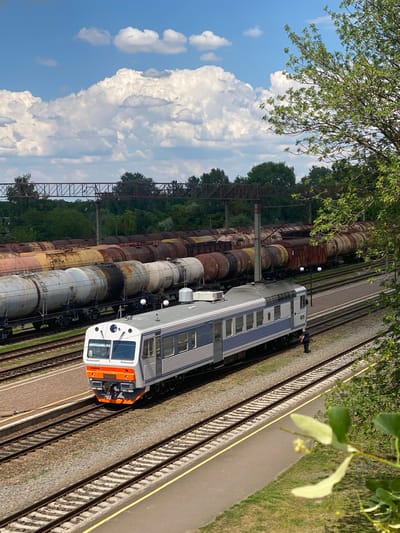 The photograph captures a sunny day at a train station in Romodan, Ukraine. In the foreground, a sleek, modern passenger train with a white and blue livery sits on a track. The train's front is orange and white, with large windows. A man in dark clothing stands beside the train. Behind the passenger train, a long line of older, weathered tank cars, some appearing rusted, stretch into the background, parked on parallel tracks. The tracks themselves are made of ballast and are well-maintained. Beyond the train cars, dense green trees line the horizon, suggesting a rural or semi-rural setting. Overhead, a bright blue sky is dotted with fluffy white clouds. A section of what appears to be an overhead railway bridge or support structure is visible in the middle ground. Lush green foliage from a tree in the foreground partially obscures the upper right corner of the image, adding a natural element to the industrial scene. The overall impression is of a working railway environment under a clear, pleasant sky.