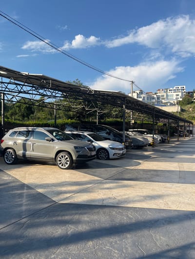 The image shows a row of parked cars under a covered parking structure. The parking structure has a metal frame and a roof, likely for shade or shelter. Several cars are visible, including a grey SUV in the foreground. The ground in the parking area is paved with patterned concrete. In the background, there are trees and modern white buildings on a hillside, consistent with the location in Krimovice, Montenegro. The sky is blue with scattered white clouds, and the sun is casting shadows, indicating it is daytime. There are no people visible in the image.