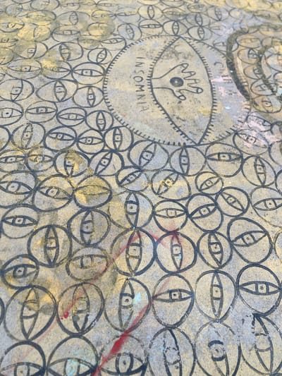 The image shows a close-up, angled view of a textured, light-colored surface with a repeated pattern of stylized eyes drawn in black. The eyes are enclosed within circles and ovals, and some have a distinct pupil and eyelid shape. There are also hints of gold and red color present, possibly from paint or staining, which creates a weathered or artistic effect. In the upper right portion of the image, there is a larger, more complex drawing that appears to be a symbolic or abstract figure, possibly an eye or a face with radiating lines, and the word "INSOMNIA" is written in capital letters in a curved line within a segmented circle. This artwork is located on a wall in Badalona, Spain, specifically on a section of the exterior wall of the Mercat Municipal del Centre, a public market. The overall impression is one of urban art or graffiti integrated into the city's infrastructure.