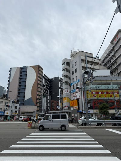 The image is rotated 90 degrees clockwise, depicting a daytime urban street scene in Osaka, Japan, under an overcast sky. The photograph captures a multi-story city block with various commercial establishments and apartment buildings, alongside a busy street with a prominent pedestrian crosswalk.

In the foreground and midground, several vehicles are visible. A white kei van is parked on the side of the road, partially on the white stripes of the crosswalk. Another white van and a darker car are seen further down the street. A traffic light pole with multiple signal heads stands near the crosswalk. A yellow and red checkered construction barrel is also present on the sidewalk.

The buildings lining the street are densely packed, featuring a mix of architectural styles. One prominent building on the left has a distinctive facade with curved elements and geometric patterns in shades of brown, white, and grey. Many buildings display colorful, multi-lingual signage, primarily in Japanese.

Visible text includes signs for "LAWSON" (a convenience store), "鳥貴族 KIZOKU 駅前店" (Torikizoku, a yakitori restaurant chain, "station front branch"), and "Conner Coffee." Another large sign advertises "ラティス niima ヨガ・ピラティス" (Lattice Niima Yoga/Pilates). Below this, an advertisement details services like "全身ほぐし 60分 3600円!" (full body massage 60 minutes 3600 yen!), "肩こり! 足ツボ!パソコンの疲れ!" (stiff shoulders! foot reflexology! computer fatigue!), with contact information and operating hours (12 PM to 12 AM). There is also a sign for "コインランドリー" (coin laundry). The sky is a uniform light grey, indicating an overcast day. The scene is typical of a bustling urban environment with commercial activity.