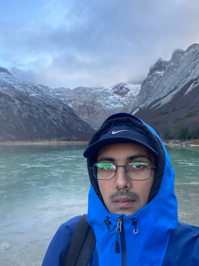 A person wearing a blue hooded jacket and glasses is facing the camera. They are also wearing a dark cap with a white Nike logo. In the background is a body of water and a mountain range, partially obscured by clouds. The scene appears to be outdoors. Location is in the city of Unknown, Argentina.