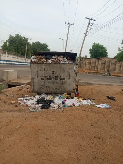 A large, grey, metal garbage bin overflowing with trash is located on a dirt road in Maiduguri, Nigeria. The bin is labeled with the words "The Presidency Ecological Fund Office" and the coat of arms of Nigeria. It is partially obscured by a small tree in the foreground, and surrounded by a substantial amount of litter. The bin is located in front of a brick wall. There are electrical poles and power lines in the background. The location is on a dirt road in the city, between a brick wall and a paved road. There are no people in the image.