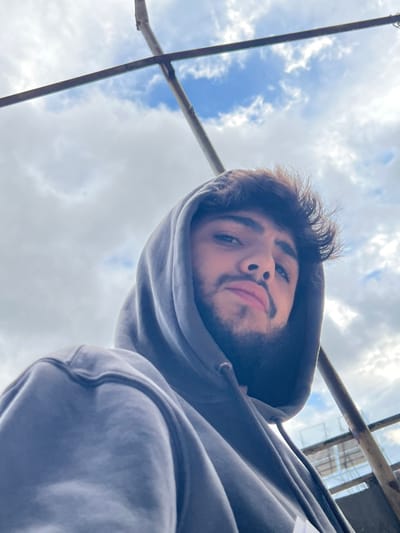 A young adult male, believed to be in Joubar Municipality, Syria, is depicted in a low-angle, outdoor portrait. He possesses dark hair and a beard, and is wearing a dark grey hooded sweatshirt with the hood raised. His gaze is directed forward, towards the camera. The background consists of a partly cloudy sky with discernible blue patches. Two dark-colored, linear structural elements, possibly metal bars or scaffolding, intersect in the upper left quadrant of the frame. A faint, lighter-colored structure, potentially a railing, is visible behind the subject's left shoulder. The overall lighting indicates daytime.