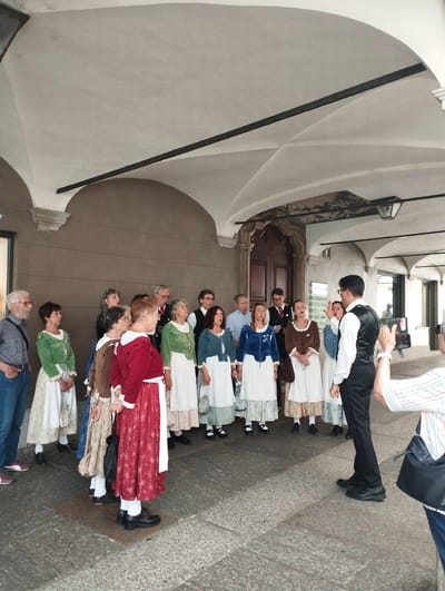 The image depicts a group of people, likely a choir, standing in an outdoor covered walkway with arched ceilings in Lugano, Switzerland. A man in a white shirt and black vest, presumably the conductor, is facing away from the camera, gesturing with his right hand as if leading the performance. A line of individuals, mostly women dressed in traditional clothing with long skirts and blouses, stand facing him, some with their mouths open as if singing. One man with glasses, wearing a white shirt and a red vest, is also part of the group. Another man in the foreground, wearing a plaid shirt and jeans, is looking at the performers. The walkway features patterned stone flooring and a large wooden door is visible in the background. The architecture suggests a historical or cultural site within Lugano.