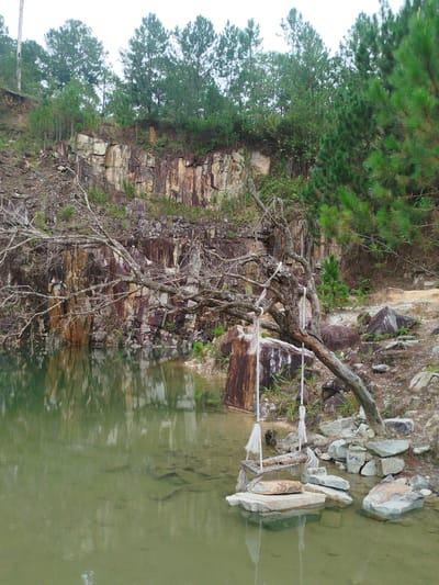 The image depicts a quarry in Lán Tranh, Vietnam, featuring a tranquil body of water, likely a quarry pit, with clear, greenish water reflecting the surrounding rock formations and trees. The quarry walls are layered with reddish-brown and grey rock, interspersed with patches of green vegetation. A fallen, dry tree branch extends over the water, from which a rustic swing is suspended. The swing consists of a wooden plank seat and two ropes tied to the branch, with fringed ends. Several large rocks are scattered around the edge of the water, and the bank is covered in smaller stones and sparse vegetation. In the background, a dense line of pine trees and other foliage crowns the quarry. The sky above is overcast, contributing to the muted light of the scene. There are no people or animals visible in the image. The overall impression is one of a natural, somewhat wild, and serene landscape transformed by past industrial activity.