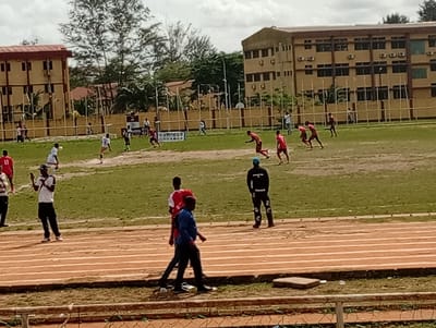 A soccer game is in progress on a field in Ijanikin, Nigeria. Several players in red jerseys are running on the grass field, while others in white are defending. A few spectators are standing on the side of the field near a track marked with white lines. Buildings are visible in the background. The sky is overcast.