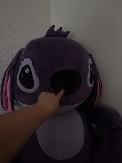 A close-up, angled shot features a large, plush purple animal, possibly a stylized bunny or bear-like creature, with one arm of a person pointing to its face. The creature is primarily purple with pink accents on the ears and lighter purple details on the face and belly. The lighting is somewhat dim, creating shadows on parts of the stuffed animal and the person's arm. The background is a neutral wall, slightly out of focus. The arm appears pale in the dim lighting, directing the viewer's attention to the face of the plush toy. Kuklen, Bulgaria is not apparent from this image.