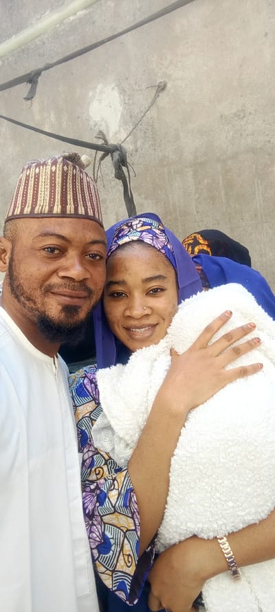 A man, a woman, and an infant are depicted in a close-up, selfie-style photograph taken in Dan Maliki, Nigeria, against a light grey concrete wall. The adult male, positioned on the left, has a dark beard and wears a white, collared garment and a patterned brown, gold, and white cap. The adult female, positioned on the right, wears a blue head covering with a visible colorful patterned band (purple, blue, gold, white) and a small silver nose stud in her right nostril. Her fingernails show a reddish brown stain. She wears a rose gold link bracelet on her left wrist. She is holding an infant, largely obscured, wrapped in a white, fluffy textile blanket. Both adults are looking towards the camera with smiles. The background wall exhibits texture, discoloration, and small white patches. Two dark, horizontal cables or ropes are visible across the upper portion of the wall, with a bundle of similar material tied below the upper cable on the right side. A light green pipe runs horizontally along the top edge of the frame. A separate rope is knotted into the wall on the upper right.