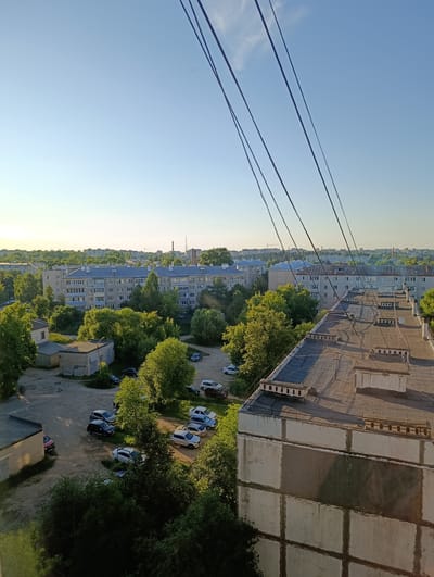 This is a high-angle, outdoor photograph taken on a clear day, likely in the early morning or late afternoon due to the warm, soft sunlight illuminating the scene. The image captures a residential area, characteristic of Alexandrov, Russia.
The main subjects are several multi-story apartment buildings, typical Soviet-era block housing, with grey tiled roofs and light-colored facades. In the foreground, the roof of a closer building is visible, featuring a weathered tar-like surface, ventilation shafts, and some debris. Several power lines or cables stretch diagonally across the sky, creating a stark visual element against the clear blue.
Surrounding the buildings is a landscape of lush green trees, their foliage dense and vibrant. The ground level reveals a dusty, unpaved area that serves as a parking lot for numerous cars. These vehicles vary in color and type, suggesting a typical urban or suburban setting. Smaller, single-story structures, possibly garages or utility buildings, are also present among the trees.
The overall impression is one of a quiet, everyday scene in a Russian residential district. The clear weather and warm light create a serene atmosphere. There is no visible text in the image.