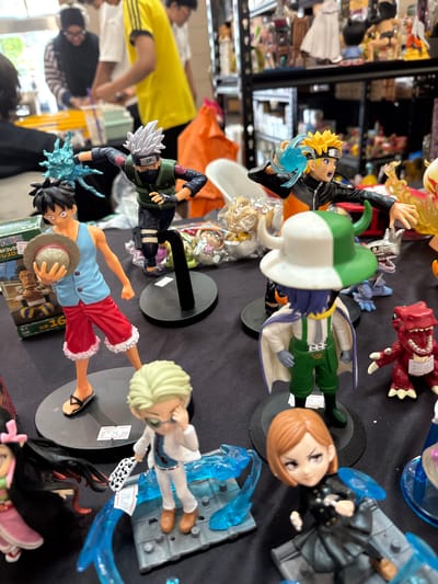 In a marketplace stall in Kuala Lumpur, Malaysia, a collection of anime figurines are displayed on a black tablecloth. In the foreground, several figures are arranged, including a character with blonde hair and a white and green hat, a character in a white suit holding a checkered flag, and a female character in a black outfit with short brown hair. Behind them, a character in red pants and a blue sleeveless shirt holds a straw hat. To the right, a red dinosaur-like figurine with a tag displaying "B 10.00" is visible. In the background, out-of-focus people are browsing through merchandise. The overall scene suggests a convention or a retail environment focused on collectible toys.