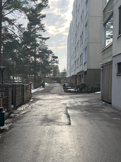 The scene depicts an outdoor pathway in Espoo, Finland, likely during late winter or early spring. In the foreground, a wet, dark asphalt surface with visible cracks occupies the majority of the frame, indicating recent moisture or melting ice. Remnants of snow are present along the left edge of the asphalt and under vegetation.

To the left, a dark gray streetlamp is partially visible. Further back on the left, a horizontal slatted wooden fence defines an area, possibly a private yard or utility enclosure. Beyond the fence, a dense line of tall evergreen trees, primarily pines, runs parallel to the pathway, forming a wooded area that extends into the background. A single conical evergreen shrub stands on a brick-paved pedestrian path that leads into the distance.

To the right, a large, multi-story residential building with a light-colored (off-white/pale green) facade and numerous rectangular windows extends into the background. The building features recessed ground-level entrances and some balcony elements. Adjacent to it in the foreground is a smaller structure with vertical wooden paneling, possibly a garage or utility access, featuring a wall-mounted light fixture and a visible address number `6B`. Low, bare shrubs line the base of the buildings. The sky above is overcast with bright glare from the sun, which is low in the sky and located towards the background. No individuals are present.