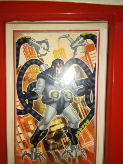 This image, captured in Ciputri, Indonesia, displays a framed, brightly illuminated comic book or animated style illustration. The central subject is a male figure, depicted with long dark hair, a pale face, and wearing red-tinted glasses. He is dressed in a light grey technological suit with prominent black and dark grey accents, featuring two large yellow-green circular components on the chest and smaller similar elements elsewhere. Four segmented mechanical tentacles, primarily black with grey stripes and some lime green circular accents, emerge from his back and lower legs. Each tentacle terminates in a three-pronged metallic claw. The figure stands in a dynamic, slightly stooped pose with his arms bent at the elbows, hands gloved in black. Two upper tentacles extend upwards, while two lower tentacles connect to the figure's black boots, forming a stable stance. The background of the illustration features a stylized cityscape composed of numerous orange-brown skyscrapers under a lighter, unidentifiable sky. The illustration is contained within a white frame, which is encased by a wider, glossy red frame.