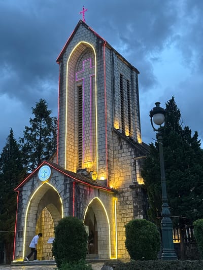 The image shows a stone church illuminated with neon lights in Sa Pa Ward, Vietnam. The church has a tall bell tower with a cross on top. The tower is decorated with a purple neon cross, and yellow neon lights outline its shape. Other parts of the church have red and yellow neon light details. A streetlight is visible to the right of the church's tower. A person in a white shirt and black pants is walking on the right side of the church. The sky is a dark shade of blue, indicating that the photo was taken in the evening.