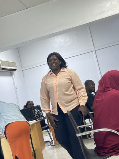 The scene depicts an indoor gathering of several individuals in a brightly lit room. The location is specified as the City of Zaria, Nigeria.
A dark-complexioned adult female, with dark hair, stands prominently in the mid-ground, slightly off-center to the right. She is wearing a light peach-colored long-sleeved button-up shirt with a ruffled collar and dark trousers. Her expression is a broad smile, and she is looking towards the left side of the frame. Her right hand rests on the back of a black office chair.
To her left, partially obscured by a light blue denim jacket and an orange skirt worn by a person in the immediate foreground, a dark-complexioned female is seated at a light brown wooden desk. This seated female, wearing a black and white striped shirt, is also smiling and looking towards the left, with a black computer monitor visible on the desk in front of her.
To the right of the central standing figure, a dark-complexioned male with short hair is partially visible, seated in a dark shirt, also looking towards the left. In the extreme foreground on the right, the back and right side of a person wearing a maroon head covering and garment are visible, seated on a black chair.
The room features plain white walls, a white ceiling with visible trim, and a light-colored tiled floor. A white wall-mounted air conditioning unit is present on the upper left wall. Several black office chairs with metal frames are visible throughout the space. The overall environment suggests an office, classroom, or meeting room setting.