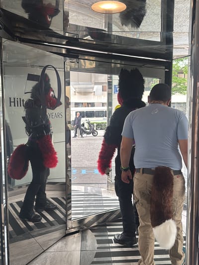 The image shows two individuals standing at the revolving doors of the Hilton Atlanta hotel, located in downtown Atlanta, Georgia, United States. Both are wearing tails, suggesting they are likely attendees of a furry convention.
On the left, one individual wears a partial fursuit consisting of a black headpiece with red accents, black clothing with red fur accents on the arms, and a large red tail. The individual is visible in the reflective surface of the revolving door, which also displays a Hilton hotel advertisement.
To the right, another person stands with their back to the camera, wearing a light blue t-shirt, khaki pants, and a brown and white tail. The person appears to be holding the door open for the individual in the fursuit.
In the background, visible through the revolving door, is a man standing near a scooter on the sidewalk outside the hotel. A white bus is also present in the background.