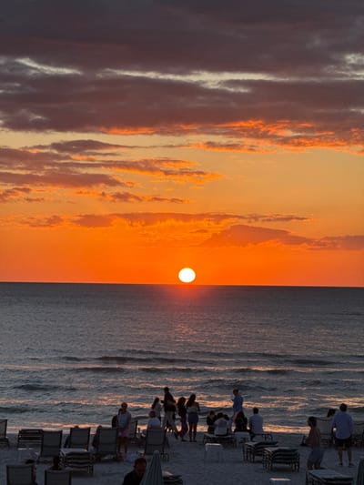 Pelican Bay, United States of America, is the location of this beach scene at sunset. A prominent, bright white sun disc is positioned low on the horizon, directly over the dark blue-grey ocean. The sky above the horizon exhibits a vibrant gradient of orange and red, transitioning to darker orange, purple, and grey hues with scattered cloud formations at higher altitudes. Gentle waves are visible near the sandy shoreline. Approximately 25 individuals are distributed across the beach. Many are silhouetted, forming small groups standing and facing the sunset, while others are seated in numerous beige-colored beach chairs arranged on the sand. Some chairs are empty or stacked. A closed beach umbrella is partially visible in the lower foreground. No specific interactions or detailed identifying features are discernible for the individuals.