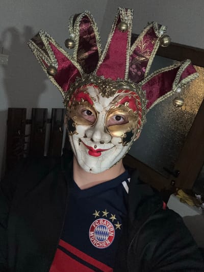 A person wearing a jester mask with red and gold accents, featuring a pointed hat with bells, is visible in the image. The mask is white with red accents around the eyes and mouth. The person is wearing a dark blue and red shirt with the logo of Bayern Munich football club. The setting appears to be indoors with a dark background. No definitive clues connect the image to the city of Goritsa, Bulgaria.