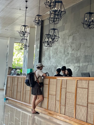 The image shows an indoor space, possibly a hotel lobby in Banyuwangi, Indonesia, based on the architecture and potential context of traveler interactions. In the center, a person wearing a cap, white t-shirt, patterned shorts, and sandals is standing at a wooden reception desk, with a backpack and what appears to be a camera bag. They seem to be interacting with two people standing behind the desk. The desk is made of a light-colored wood frame with a woven pattern filling the panels. Above, there are several black geometric cage-like light fixtures suspended from the ceiling. The ceiling is a neutral gray color with a tiled pattern. There is an apparent window with natural light and greenery visible in the background.