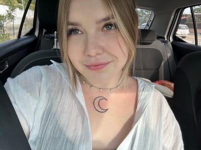 A woman with blonde hair, green eyes, and a moon tattoo on her chest is photographed from an inverted perspective inside a car. She is wearing a white blouse and a silver necklace with pearls and star-shaped accents. The car interior includes black seats and a grey dashboard. Outside the car window, there is foliage and a glimpse of a building and street. The image was possibly taken in Sutomore, Montenegro.