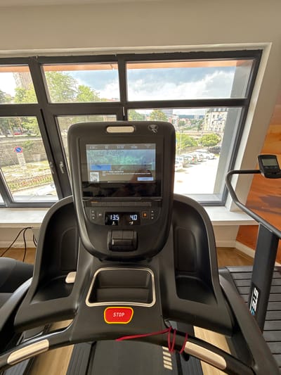 A close-up, eye-level, indoor shot shows the console and handlebars of a black PRECOR treadmill. The treadmill is positioned in front of a large, angled window that offers a view of Gabrovo, Bulgaria. Outside, trees, buildings, a river, and parked cars are visible. The console displays workout information including "Total Distance 0.58 km", "Calories 82", "Time Remaining 51:10", and "Pace 15:00". A promotional message reads "GET 1 FREE MONTH!". Below the screen, numerical readouts show "13.5" and "4.0" with icons that suggest speed and incline. A red "STOP" button is prominent on the right handlebar. The overall environment appears to be a gym or fitness center.